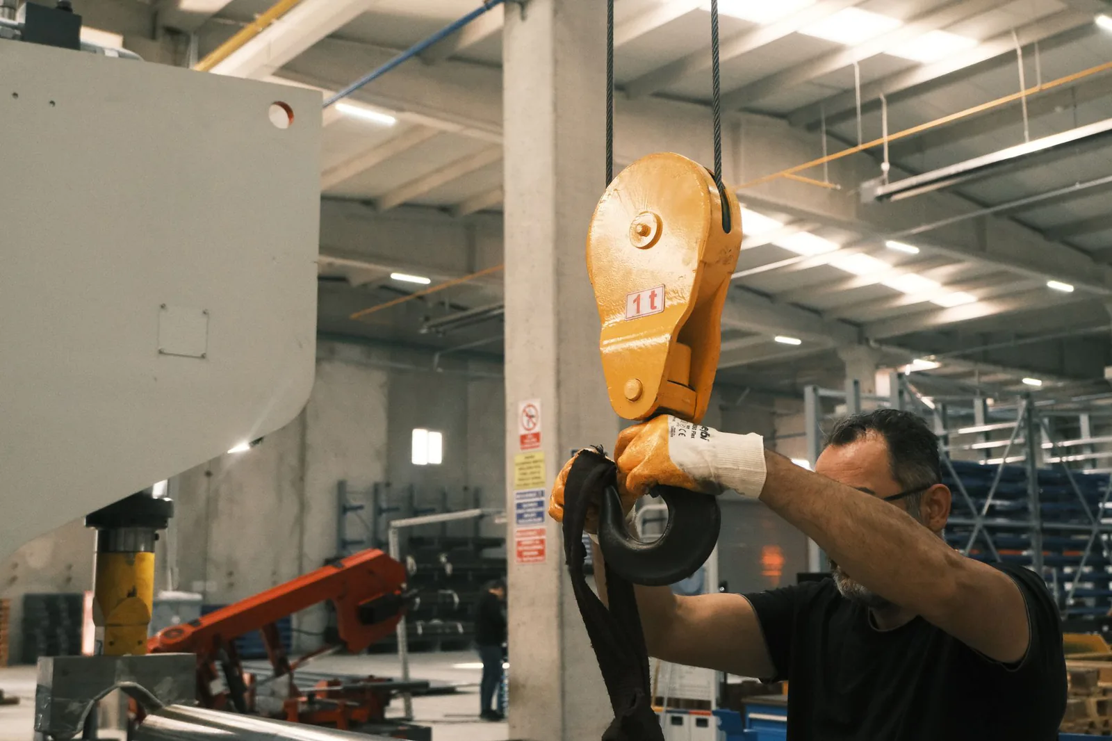 Worker in gloves fitting a lifting sling to the yellow hook block of an overhead hoist in a factory hall