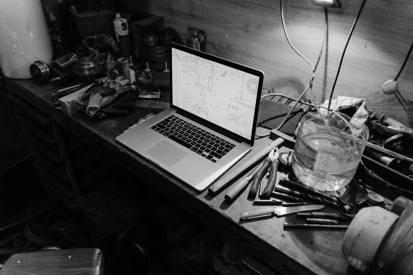 Open laptop showing technical drawings on a cluttered workbench, among pliers, hand tools and a jar of water, in black and white