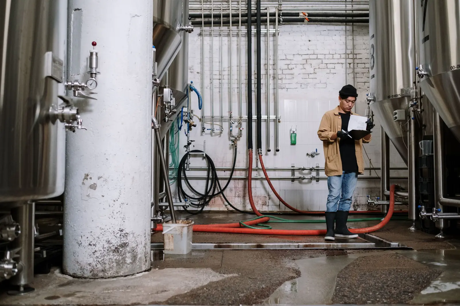Person in wellington boots holding a clipboard and pen among stainless steel tanks, hoses and pipework on a wet factory floor