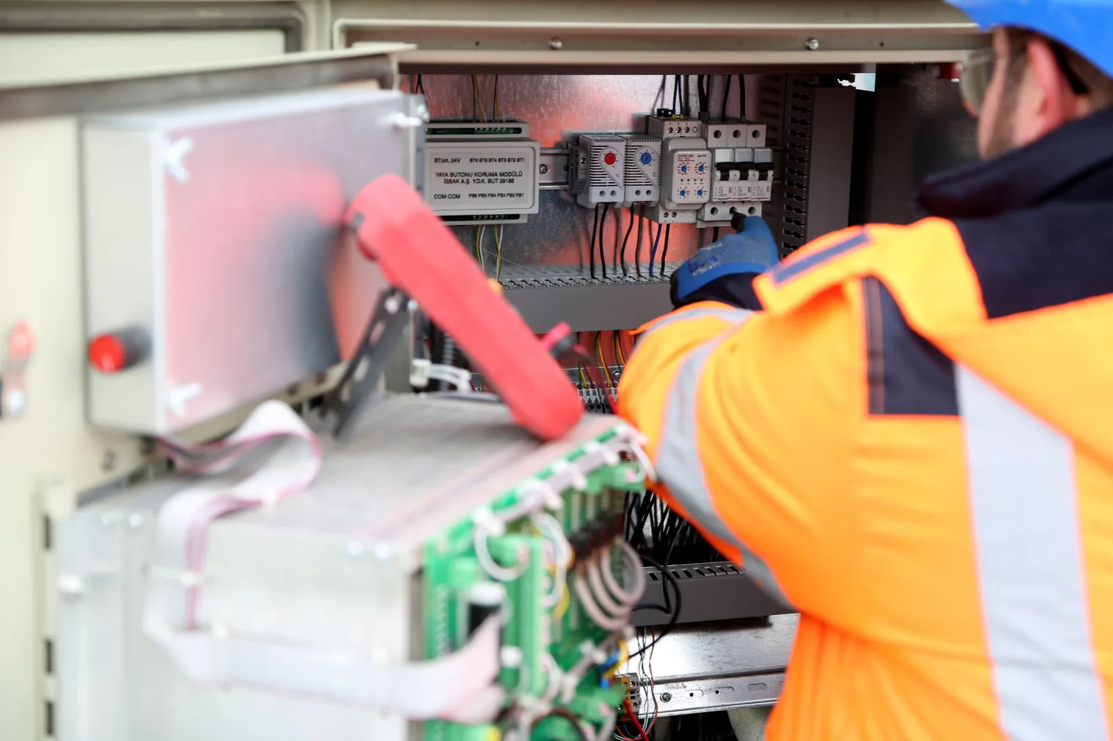 Technicien en veste orange et casque bleu qui intervient dans une armoire électrique ouverte