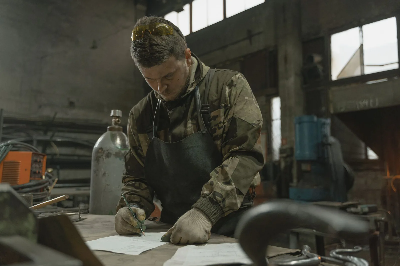 Homme en gants de travail qui écrit au stylo sur une feuille posée sur l'établi d'un atelier de métallerie