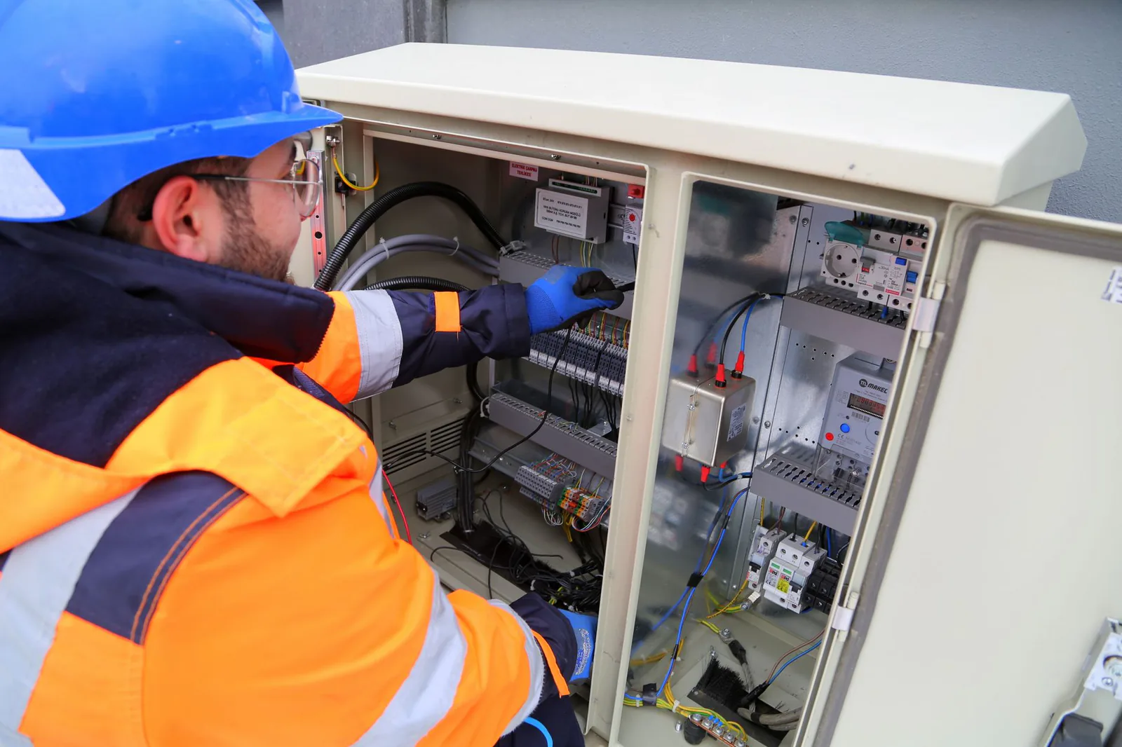 Engineer in a blue hard hat and orange high-visibility jacket working inside an open outdoor control cabinet full of wiring and breakers