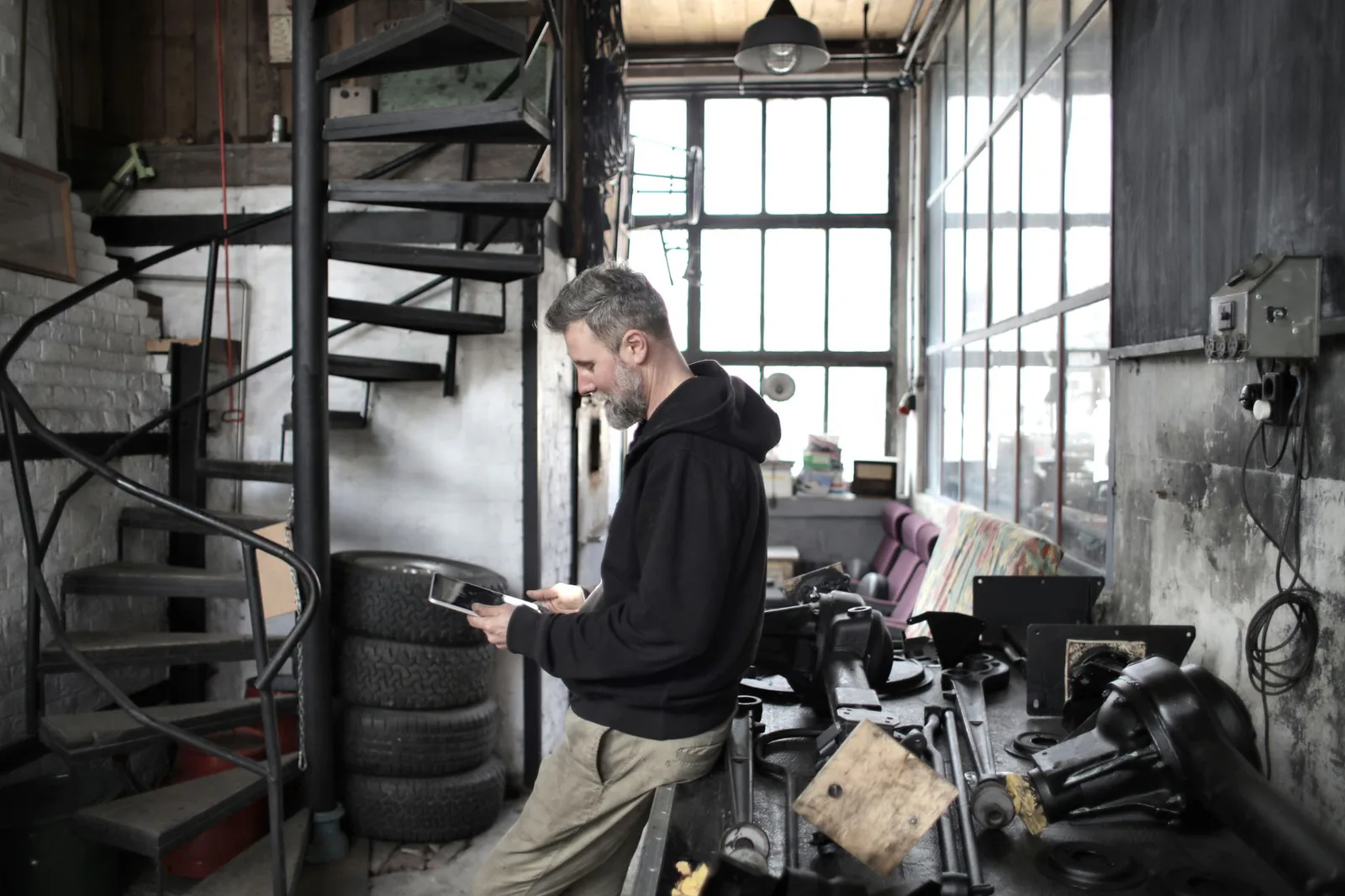 Bearded man in a dark hoodie reading a tablet beside a cluttered workbench in an old workshop with tall windows