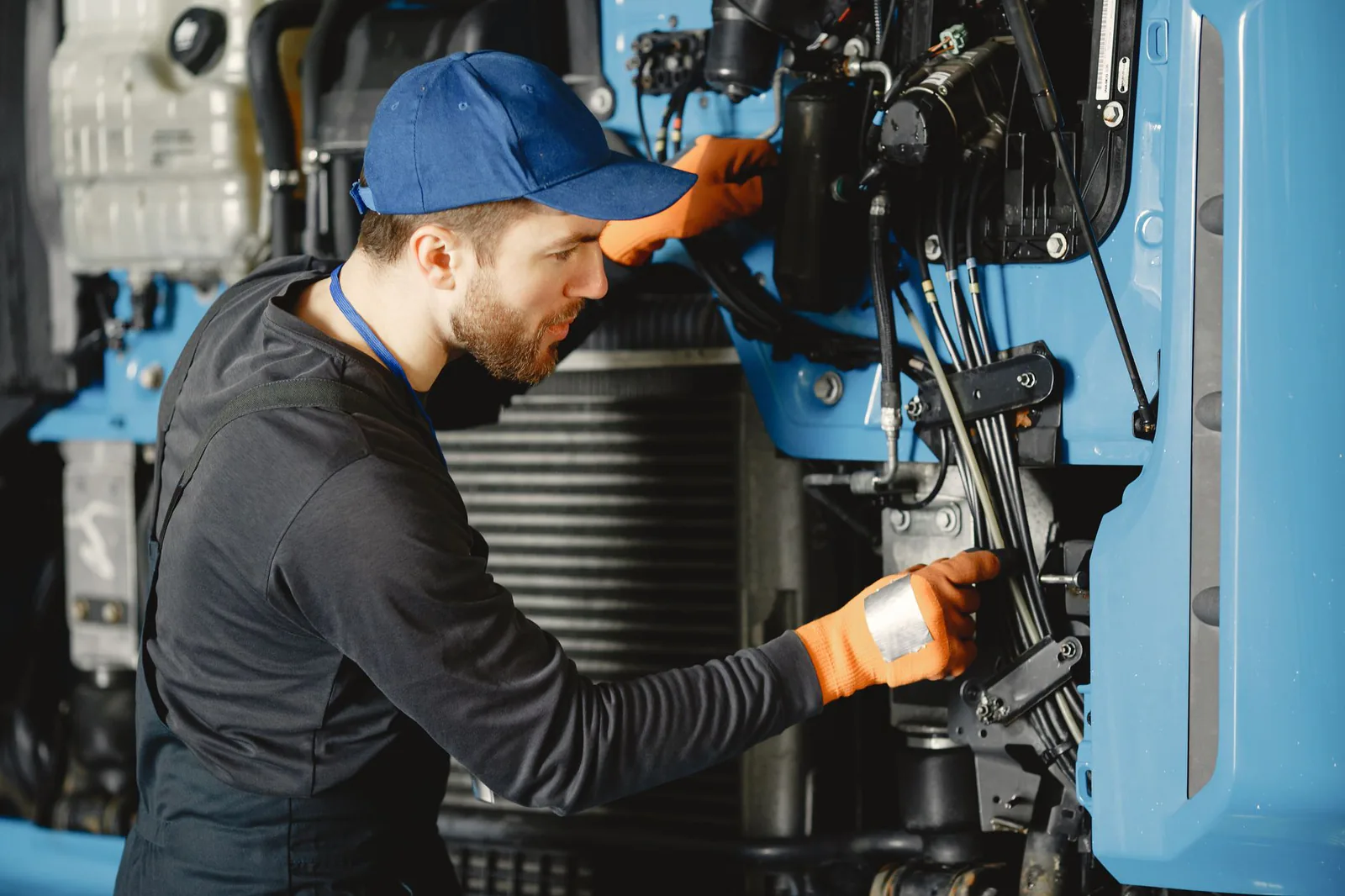 Technicien en casquette bleue et gants orange qui intervient sur le faisceau de câbles d'un engin, cabine relevée