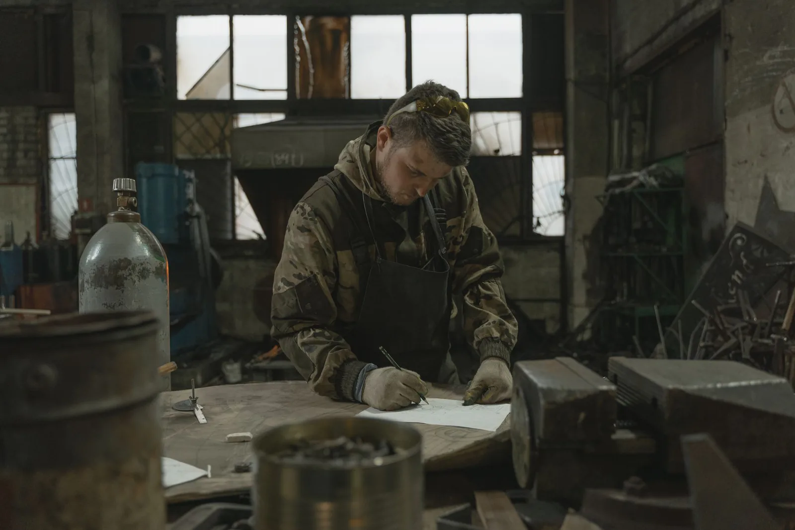 Homme en veste de travail qui écrit au stylo sur une feuille posée sur une table d'atelier, devant une grande verrière