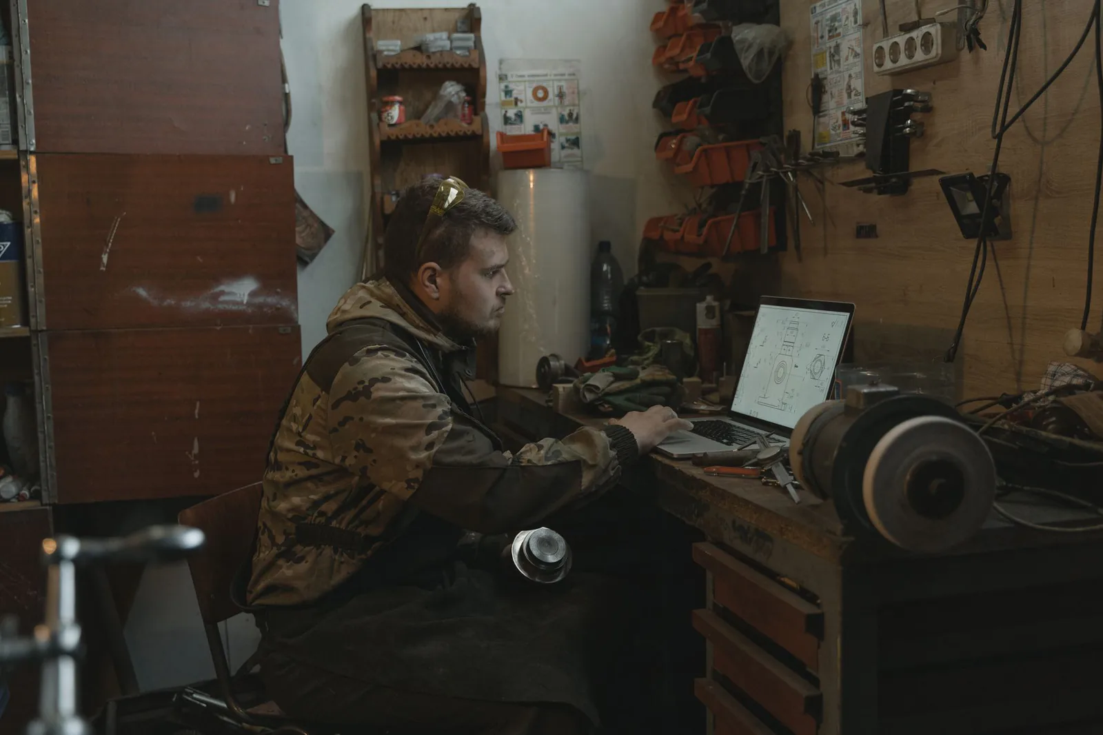Homme assis devant un ordinateur portable posé sur l'établi d'un atelier, près d'un touret à meuler et d'outils accrochés au mur