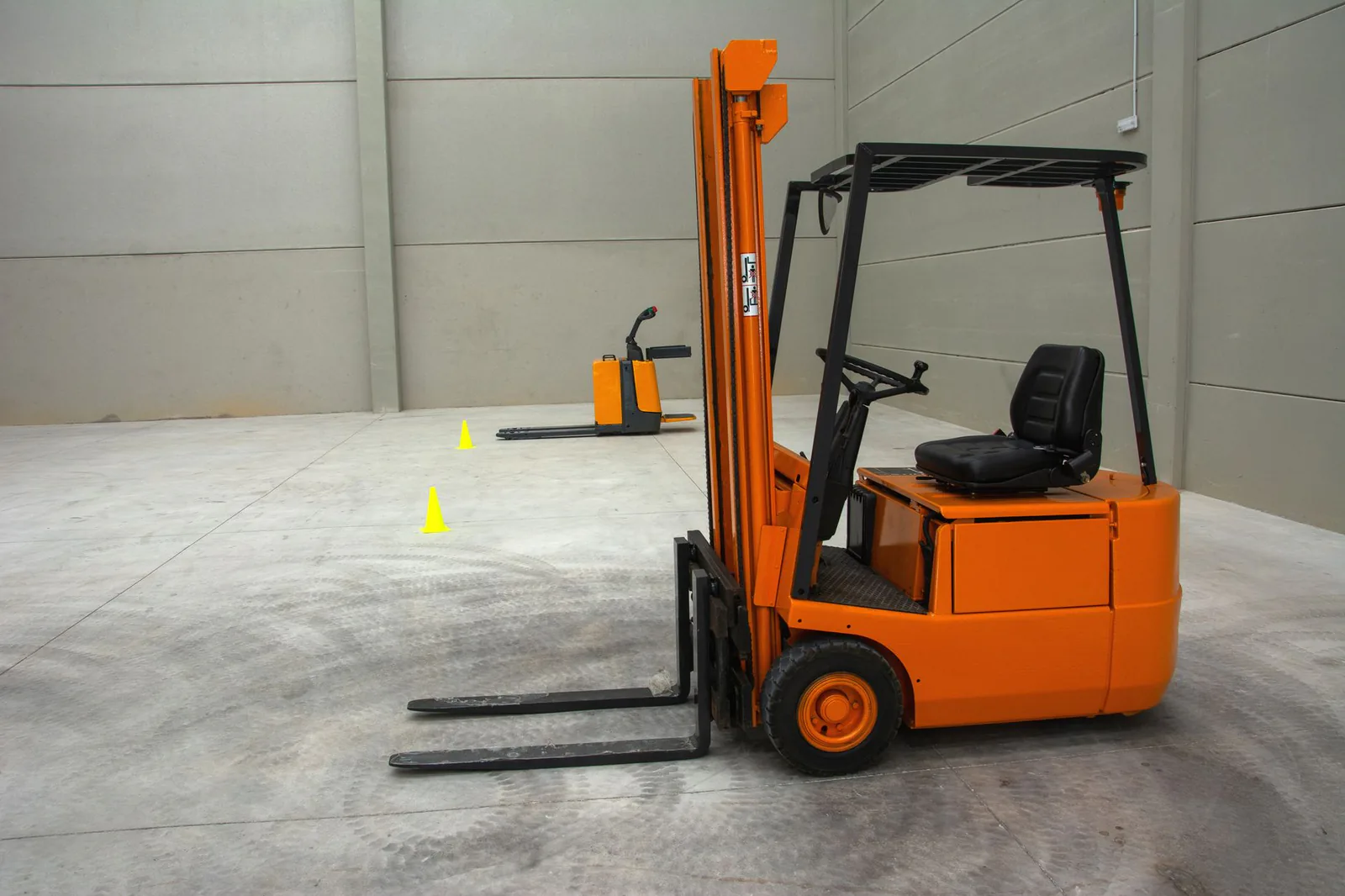 Chariot élévateur orange stationné seul dans un grand atelier au sol de béton, un transpalette en arrière-plan