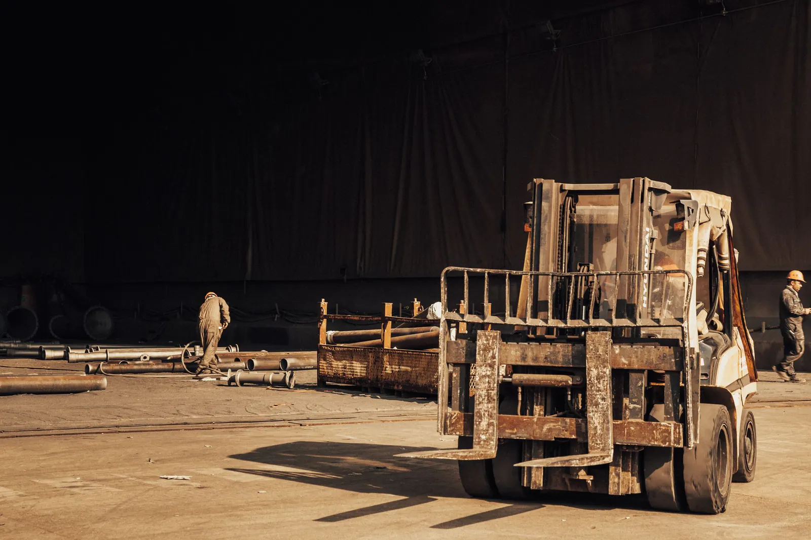 Chariot élévateur usé aux fourches rouillées dans une halle industrielle sombre, deux ouvriers parmi des tuyaux d'acier
