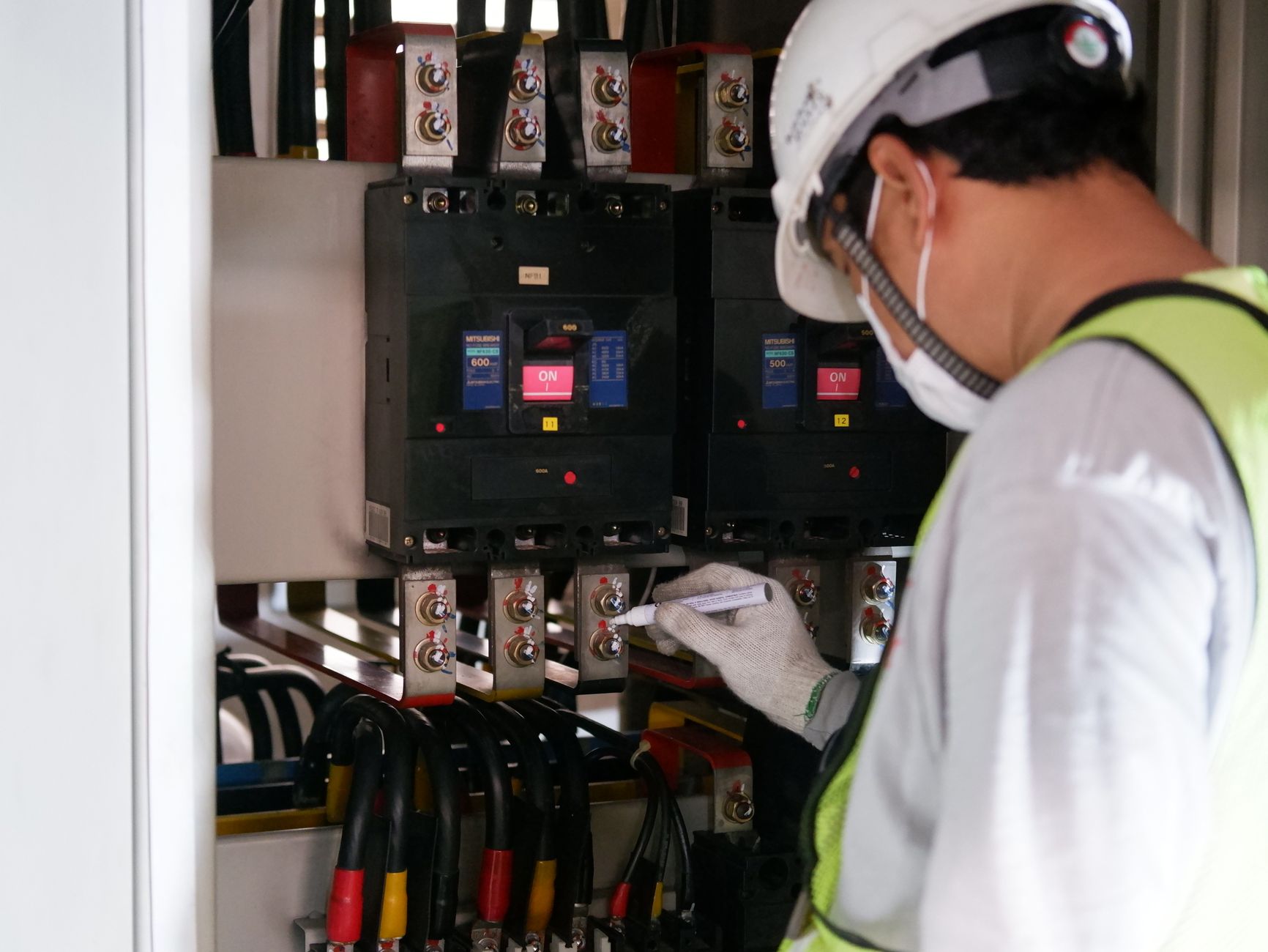Technicien casqué relevant des valeurs sur un tableau électrique, stylo à la main